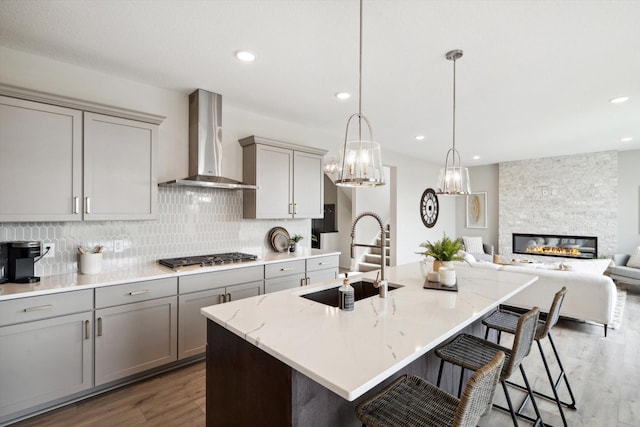 kitchen with sink, wall chimney exhaust hood, stainless steel gas cooktop, dark hardwood / wood-style floors, and an island with sink