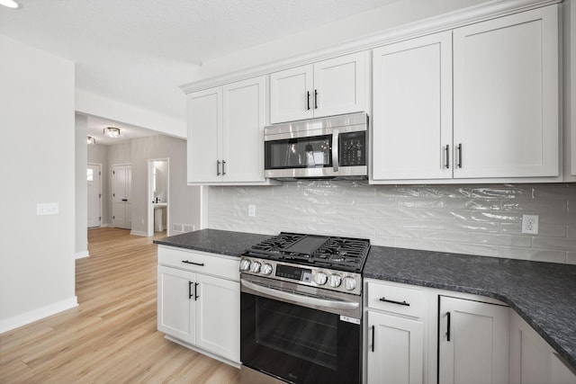 kitchen with appliances with stainless steel finishes, white cabinetry, dark stone countertops, backsplash, and light hardwood / wood-style flooring