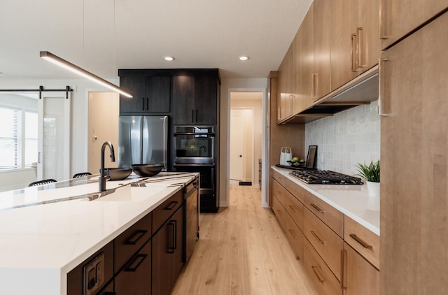 kitchen featuring appliances with stainless steel finishes, backsplash, a kitchen island with sink, a barn door, and light hardwood / wood-style floors