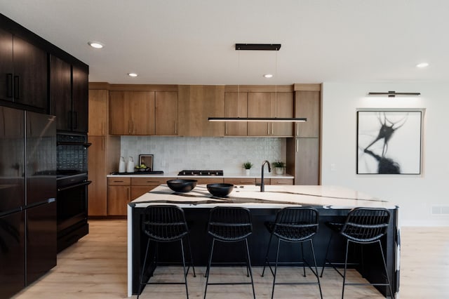 kitchen with decorative light fixtures, light wood-type flooring, an island with sink, and black appliances