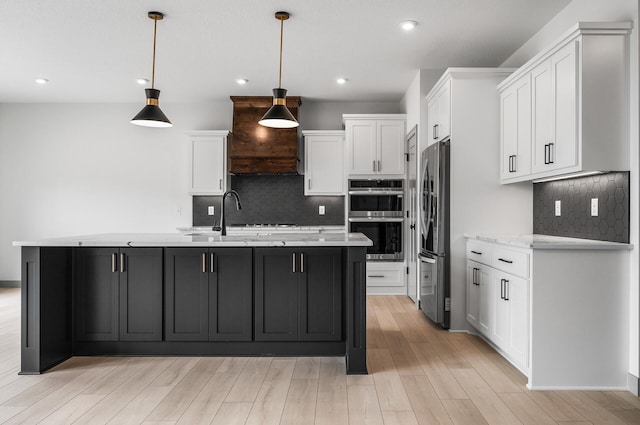 kitchen with an island with sink, appliances with stainless steel finishes, hanging light fixtures, light wood-style floors, and white cabinetry