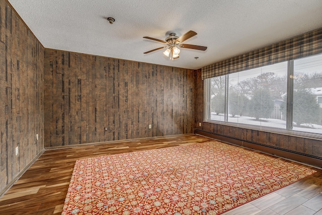 unfurnished room with ceiling fan, a textured ceiling, wood finished floors, wood walls, and baseboard heating