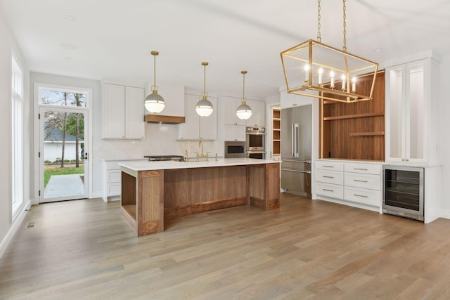 kitchen with hanging light fixtures, beverage cooler, light hardwood / wood-style flooring, an island with sink, and white cabinets