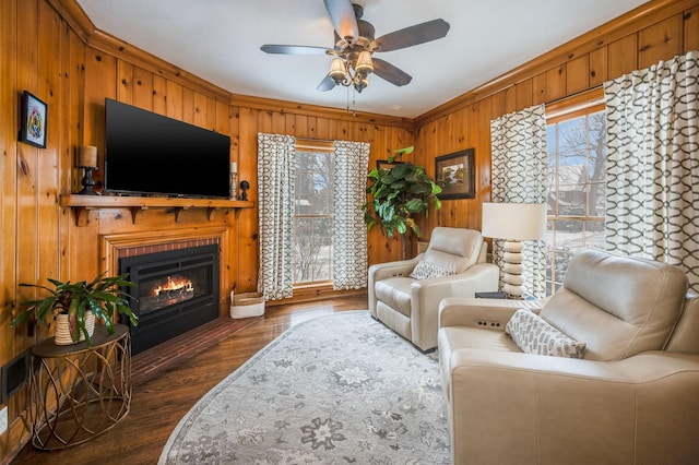 living area with wooden walls, a ceiling fan, a fireplace with flush hearth, dark wood-type flooring, and crown molding