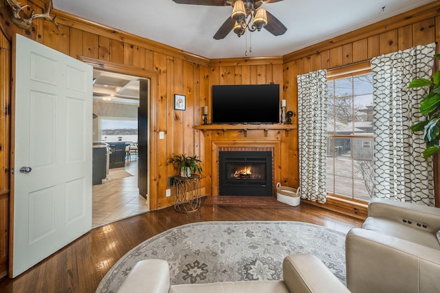 living area featuring wooden walls, crown molding, wood finished floors, a glass covered fireplace, and a ceiling fan