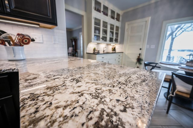 kitchen featuring dark cabinetry, light stone countertops, ornamental molding, glass insert cabinets, and backsplash