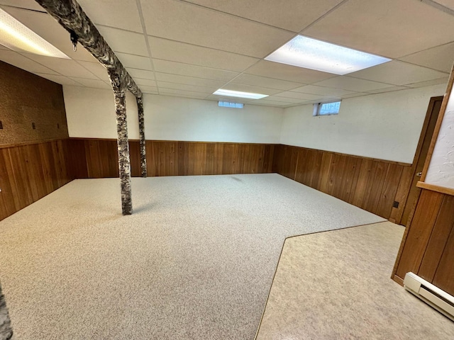 basement featuring carpet, a paneled ceiling, baseboard heating, and wooden walls