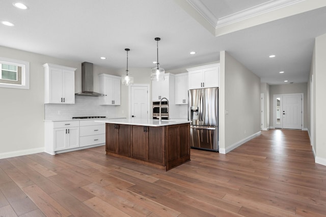 kitchen with white cabinetry, wall chimney exhaust hood, wood-type flooring, a center island with sink, and appliances with stainless steel finishes