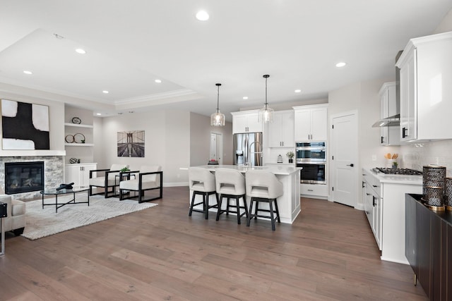 kitchen with appliances with stainless steel finishes, decorative light fixtures, white cabinets, a stone fireplace, and an island with sink