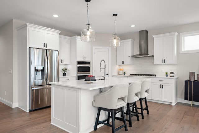 kitchen with white cabinetry, wall chimney range hood, a kitchen island with sink, appliances with stainless steel finishes, and hardwood / wood-style flooring