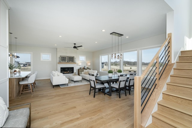 dining space featuring light wood finished floors, a fireplace, recessed lighting, stairs, and ceiling fan with notable chandelier