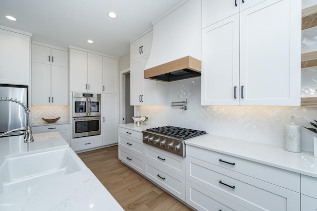 kitchen with premium range hood, backsplash, appliances with stainless steel finishes, white cabinets, and light wood finished floors