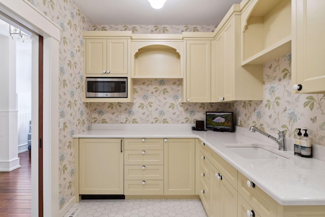 kitchen featuring stainless steel microwave, a sink, wallpapered walls, cream cabinetry, and light countertops