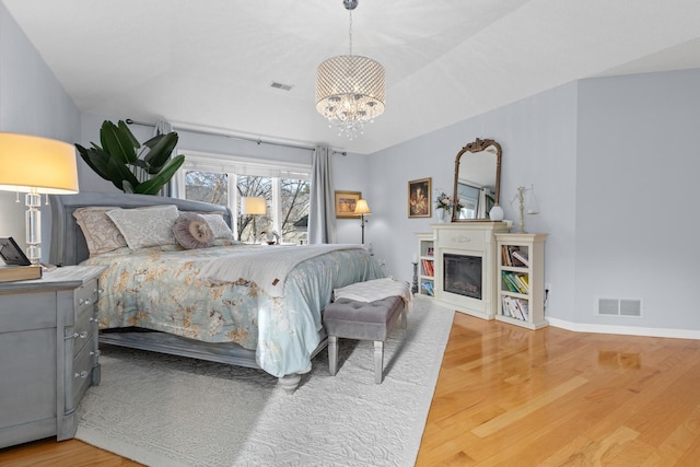 bedroom featuring baseboards, visible vents, a glass covered fireplace, wood finished floors, and an inviting chandelier