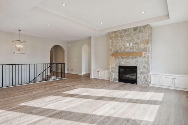 unfurnished living room with a tray ceiling, a stone fireplace, a chandelier, and light wood-type flooring