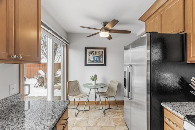 kitchen featuring light stone counters, stainless steel appliances, brown cabinetry, ceiling fan, and baseboards