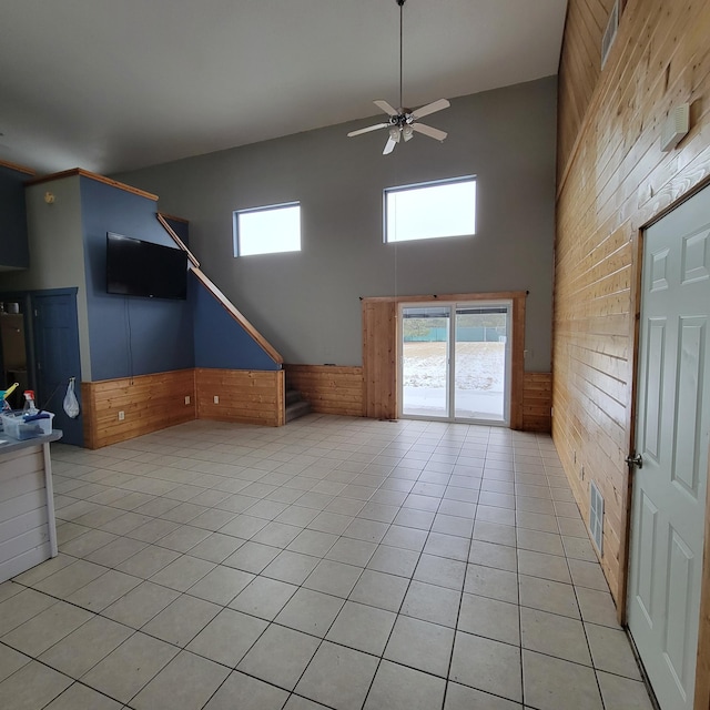 unfurnished living room with ceiling fan, wooden walls, light tile patterned floors, and a high ceiling