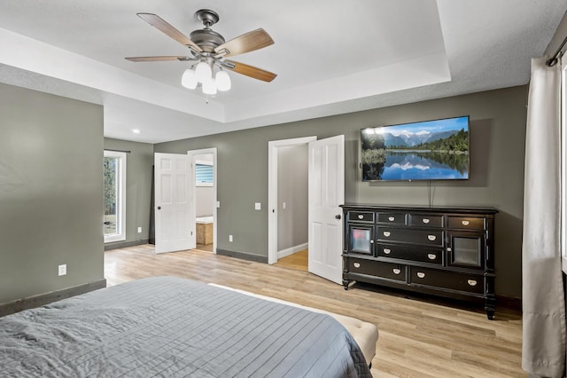 bedroom with light wood-type flooring, connected bathroom, a tray ceiling, and ceiling fan