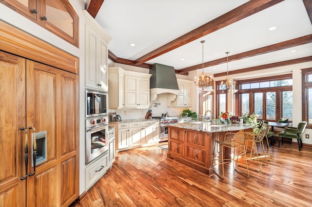 kitchen featuring light stone countertops, stainless steel appliances, a breakfast bar area, a center island with sink, and custom range hood