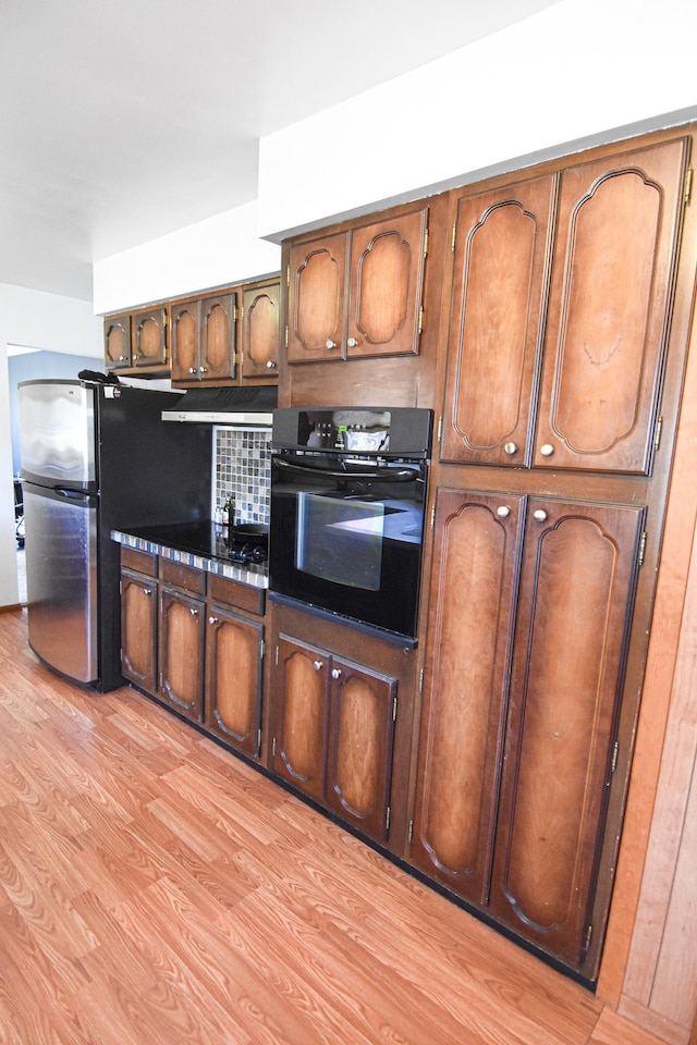 kitchen with stainless steel refrigerator, black oven, decorative backsplash, and light hardwood / wood-style floors