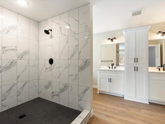 full bath with a sink, two vanities, wood finished floors, visible vents, and tiled shower