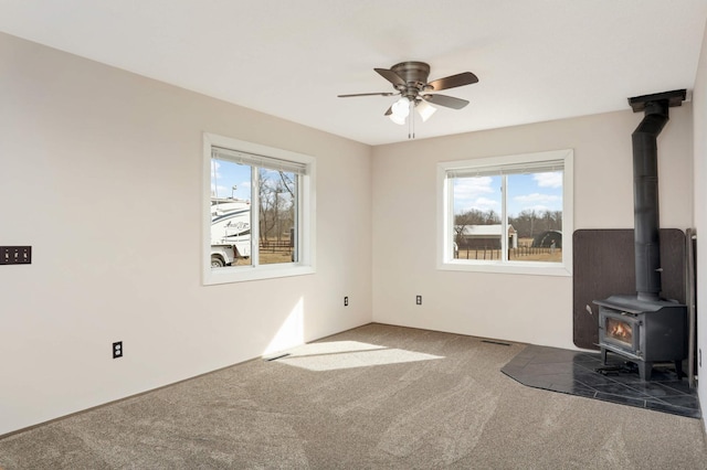 unfurnished living room featuring a wood stove, a healthy amount of sunlight, carpet floors, and ceiling fan