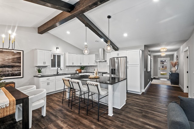 kitchen featuring wall chimney range hood, a kitchen island, lofted ceiling with beams, stainless steel fridge, and white cabinets