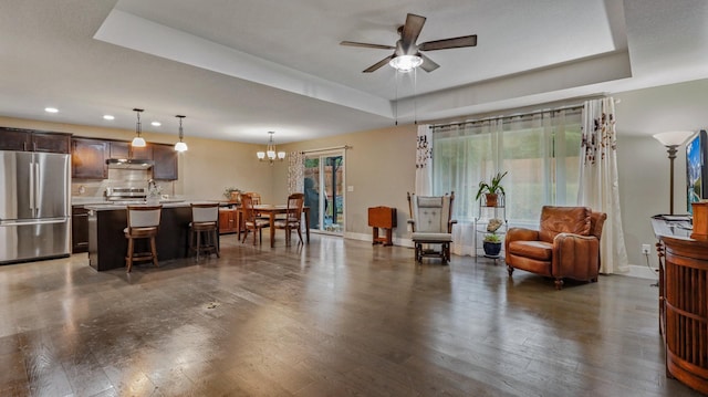 living room featuring ceiling fan with notable chandelier, a tray ceiling, and dark wood-type flooring