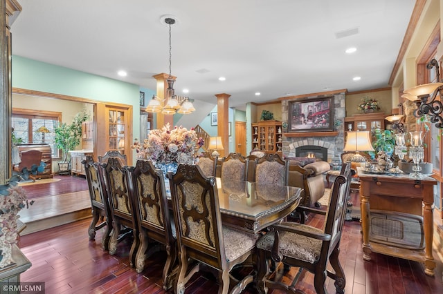 dining room featuring a stone fireplace, a chandelier, ornate columns, crown molding, and dark wood-type flooring