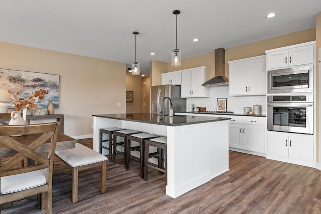 kitchen featuring appliances with stainless steel finishes, wall chimney exhaust hood, a kitchen island with sink, white cabinetry, and hanging light fixtures