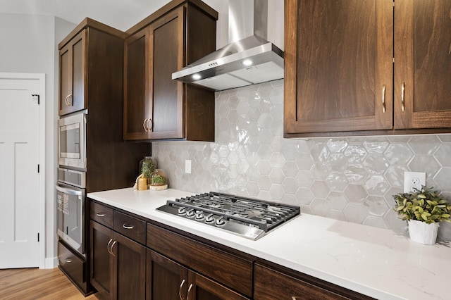 kitchen featuring decorative backsplash, dark brown cabinets, wall chimney range hood, and appliances with stainless steel finishes