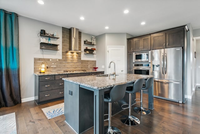 kitchen featuring dark wood-type flooring, sink, wall chimney exhaust hood, an island with sink, and appliances with stainless steel finishes