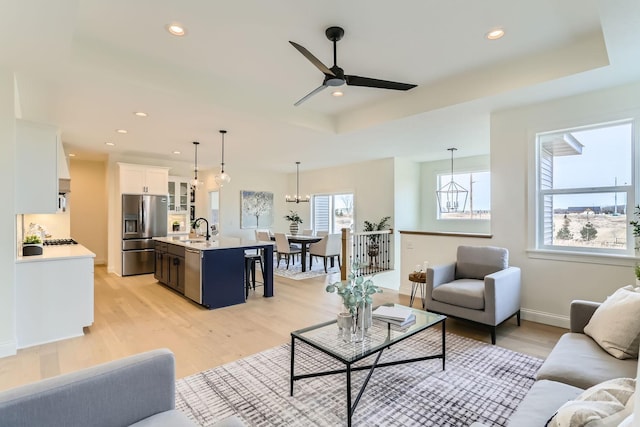 living room with ceiling fan with notable chandelier, light hardwood / wood-style floors, a raised ceiling, and sink