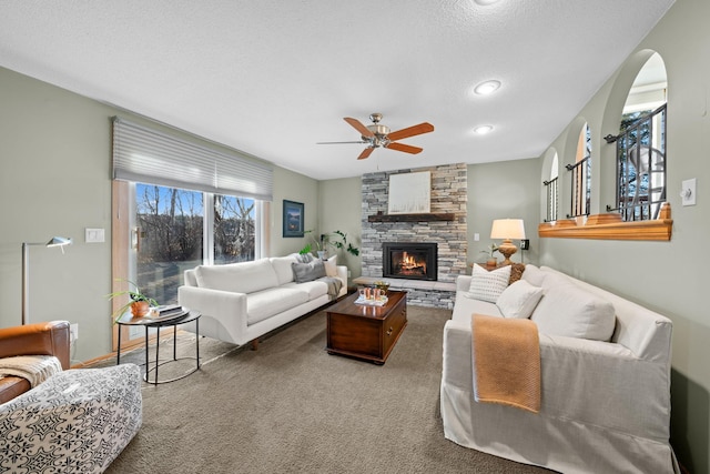 carpeted living room with a textured ceiling, ceiling fan, and a stone fireplace