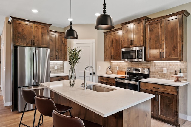 kitchen featuring appliances with stainless steel finishes, a kitchen island with sink, sink, decorative light fixtures, and light hardwood / wood-style flooring