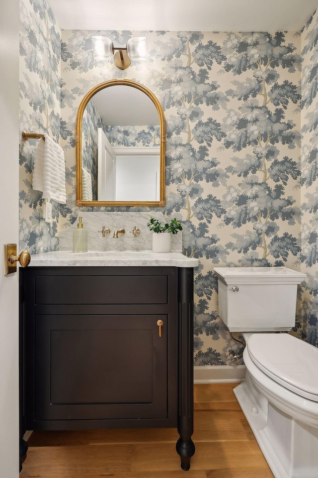 half bath with wallpapered walls, baseboards, toilet, wood finished floors, and vanity