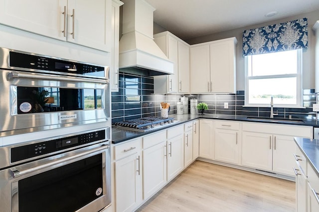 kitchen featuring sink, premium range hood, appliances with stainless steel finishes, white cabinets, and light wood-type flooring