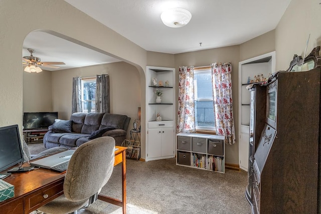 carpeted office space featuring ceiling fan, built in shelves, and plenty of natural light