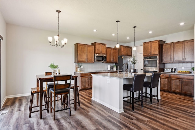 kitchen with a notable chandelier, dark wood-style floors, backsplash, a center island with sink, and appliances with stainless steel finishes