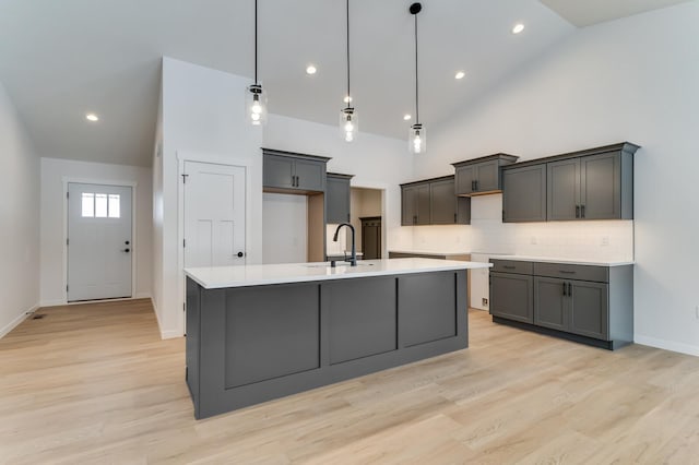 kitchen with high vaulted ceiling, a sink, light wood-style floors, decorative backsplash, and a center island with sink