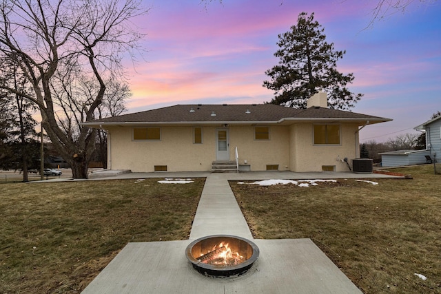 back house at dusk with central air condition unit, an outdoor fire pit, and a lawn