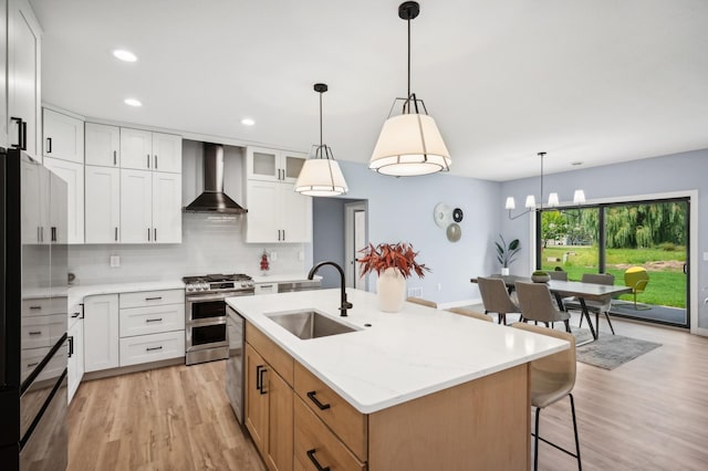 kitchen with appliances with stainless steel finishes, wall chimney exhaust hood, sink, white cabinets, and an island with sink