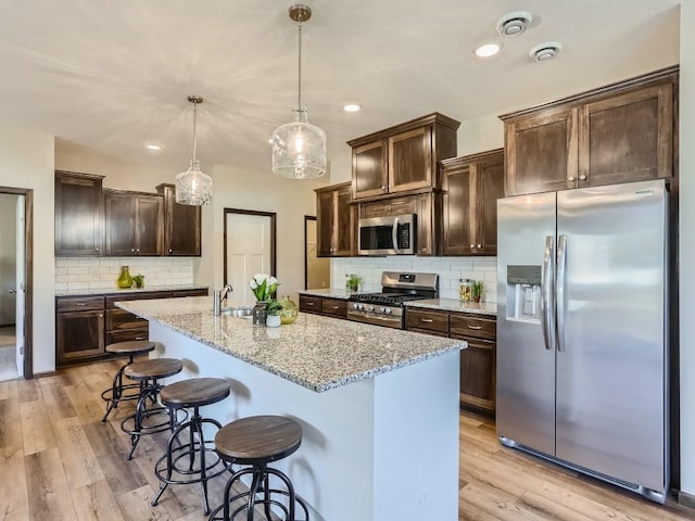 kitchen with stainless steel appliances, tasteful backsplash, light stone counters, decorative light fixtures, and a kitchen island with sink