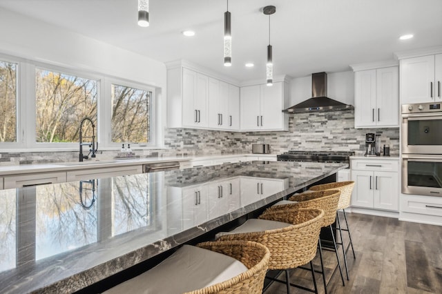 kitchen with wall chimney range hood, double oven, pendant lighting, a breakfast bar area, and white cabinets