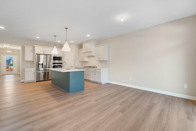 kitchen with appliances with stainless steel finishes, decorative light fixtures, white cabinets, a center island with sink, and light hardwood / wood-style flooring