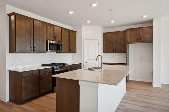 kitchen with light stone countertops, sink, stainless steel appliances, light hardwood / wood-style flooring, and an island with sink