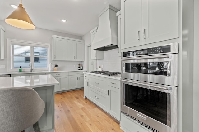 kitchen with backsplash, custom exhaust hood, stainless steel appliances, light hardwood / wood-style flooring, and hanging light fixtures