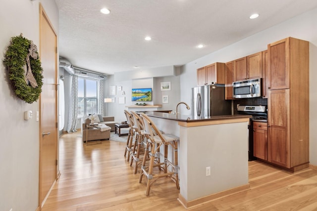 kitchen with open floor plan, a kitchen island with sink, stainless steel appliances, dark countertops, and brown cabinetry