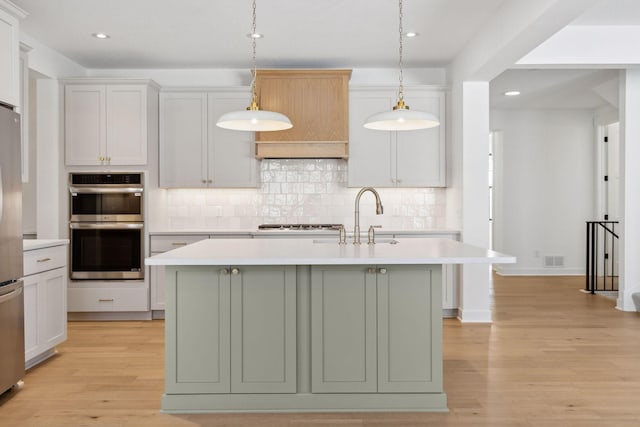 kitchen with stainless steel appliances, a center island with sink, light countertops, and decorative light fixtures