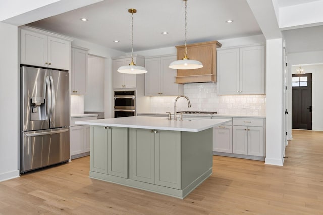 kitchen with light countertops, appliances with stainless steel finishes, light wood finished floors, an island with sink, and pendant lighting